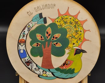 May include: A round wooden plaque with a painted design depicting a stylized tree, a bird, and a sun. The plaque is on a wooden easel and has the text "El Salvador" written on it.