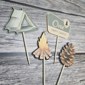 May include: Four wooden cake toppers with a camping theme. The toppers include a tent, a campfire, a pine cone, and a sign that says "One Happy Camper". The toppers are on wooden sticks.