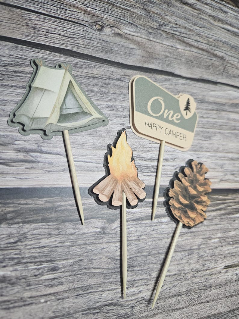 May include: Four wooden cake toppers with a camping theme. The toppers include a tent, a campfire, a pine cone, and a sign that says "One Happy Camper". The toppers are on wooden sticks.