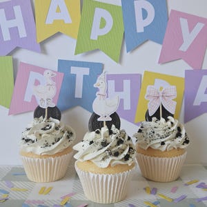 May include: Three cupcakes with white frosting and crushed chocolate cookie crumbles. Each cupcake features a decorative topper: two show white geese wearing pink accessories, and one shows a pink gingham bow, all set against a Happy Birthday banner.