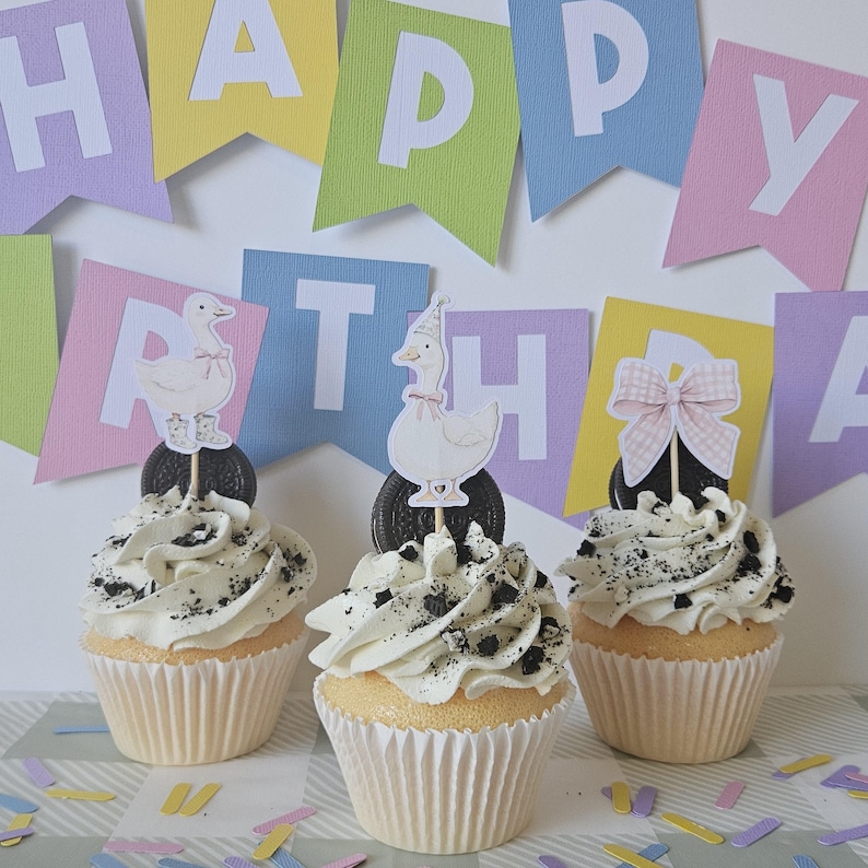 May include: Three cupcakes with white frosting and crushed chocolate cookie crumbles. Each cupcake features a decorative topper: two show white geese wearing pink accessories, and one shows a pink gingham bow, all set against a Happy Birthday banner.