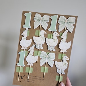 Puede incluir: Un conjunto de adornos para pasteles con tem&aacute;tica de patos sobre una tarjeta marr&oacute;n. Los adornos incluyen el n&uacute;mero "1" en verde claro, lazos y patos con sombreros y botas. La tarjeta contiene la informaci&oacute;n de contacto del vendedor.