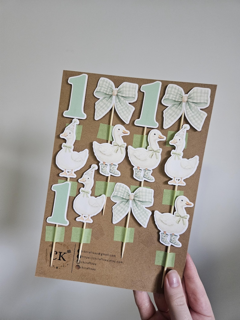 Puede incluir: Un conjunto de adornos para pasteles con tem&aacute;tica de patos sobre una tarjeta marr&oacute;n. Los adornos incluyen el n&uacute;mero "1" en verde claro, lazos y patos con sombreros y botas. La tarjeta contiene la informaci&oacute;n de contacto del vendedor.