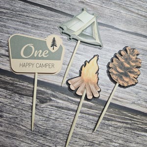 May include: Set of four camping-themed cake toppers. Includes a sign that reads "One Happy Camper", a tent, a campfire, and a pinecone. The toppers are in shades of beige, gray, and brown, with wooden sticks.