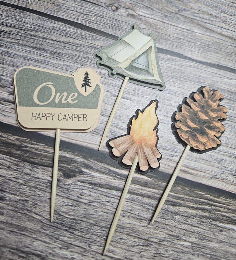 May include: Set of four camping-themed cake toppers. Includes a sign that reads "One Happy Camper", a tent, a campfire, and a pinecone. The toppers are in shades of beige, gray, and brown, with wooden sticks.