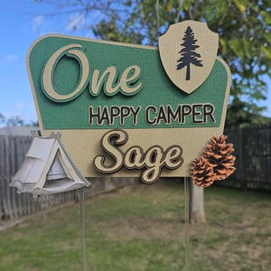 May include: A decorative sign with the words "One Happy Camper Sage" in a retro style. The sign features a tent, pine cones, and a shield with a tree. The sign is green, tan, and white.