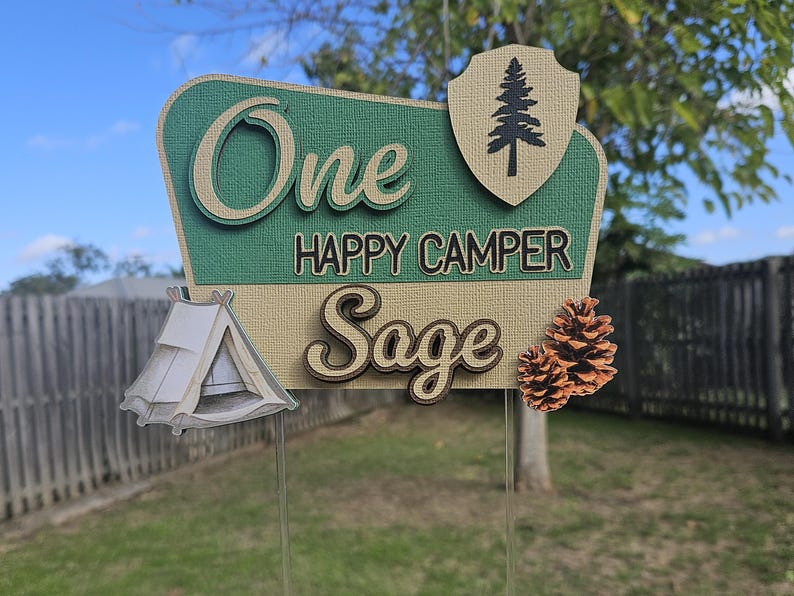 May include: A decorative sign with the words "One Happy Camper Sage" in a retro style. The sign features a tent, pine cones, and a shield with a tree. The sign is green, tan, and white.