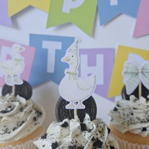 May include: Three cupcakes topped with white frosting and crushed dark cookie pieces. Each cupcake features a paper topper: a white goose wearing a floral party hat and green bow, a goose in green boots, and a large green and white checkered bow.