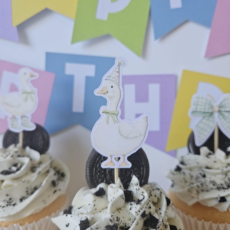 May include: Three cupcakes topped with white frosting and crushed dark cookie pieces. Each cupcake features a paper topper: a white goose wearing a floral party hat and green bow, a goose in green boots, and a large green and white checkered bow.