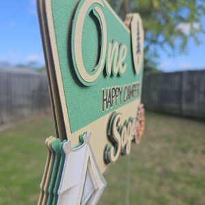 May include: A layered, green and tan sign with the words "One Happy Camper Sage" and a tent graphic. The sign is designed with a camping theme, perfect for a first birthday celebration. The sign is held by a clear plastic stick.