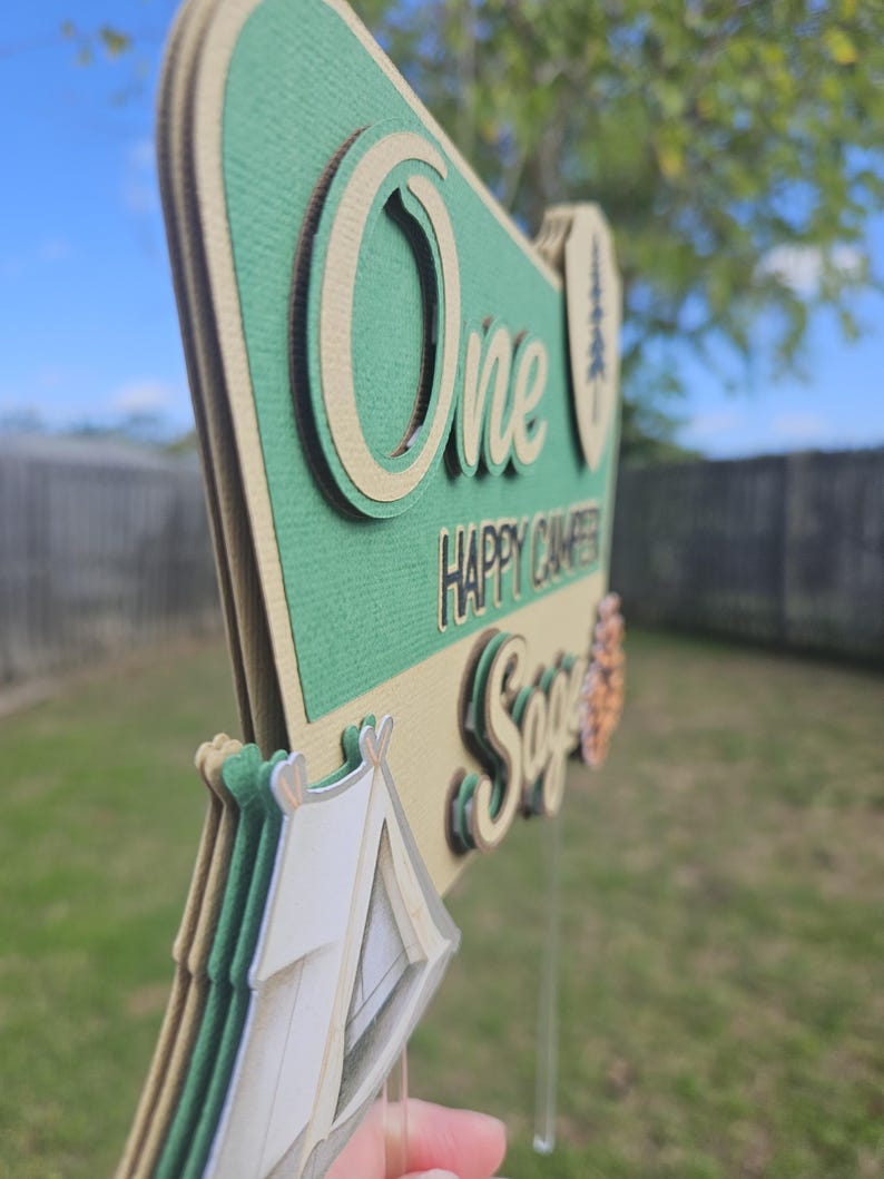 May include: A layered, green and tan sign with the words "One Happy Camper Sage" and a tent graphic. The sign is designed with a camping theme, perfect for a first birthday celebration. The sign is held by a clear plastic stick.