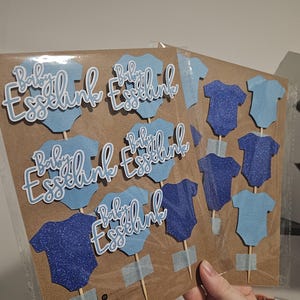 Op de afbeelding: Baby Esselink taarttoppers in lichtblauw en marineblauw. De toppers hebben de vorm van babyrompertjes en bevatten de tekst "Baby Esselink" in een sierlijk lettertype. Sommige zijn glinsterend. De toppers zijn bevestigd aan houten stokjes.