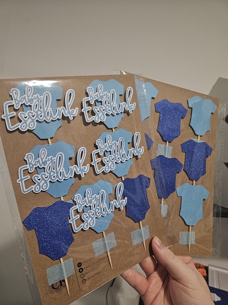 Op de afbeelding: Baby Esselink taarttoppers in lichtblauw en marineblauw. De toppers hebben de vorm van babyrompertjes en bevatten de tekst "Baby Esselink" in een sierlijk lettertype. Sommige zijn glinsterend. De toppers zijn bevestigd aan houten stokjes.