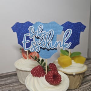 Op de afbeelding: Cupcakes met witte glazuur, versierd met vers fruit en baby-thema toppers. De toppers zijn blauwe rompertjes met de tekst "Baby Esselink" in witte letters. De cupcakes zijn versierd met aardbeien, frambozen en mango.