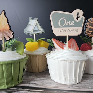 May include: Four cupcakes with camping-themed toppers. One cupcake has a campfire topper, another has a tent, a third has a sign that says "One Happy Camper", and the last has a pinecone. Each cupcake is decorated with fruit.