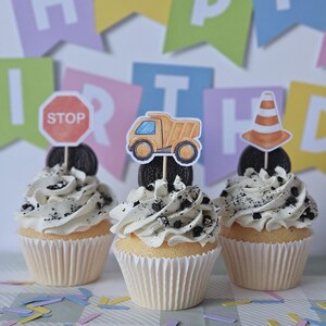 Puede incluir: Tres cupcakes de vainilla con glaseado blanco y migas de galleta de chocolate. Cada cupcake tiene un adorno de papel: una señal de stop roja, un camión volquete amarillo y un cono de tráfico naranja, montados sobre una galleta oscura.