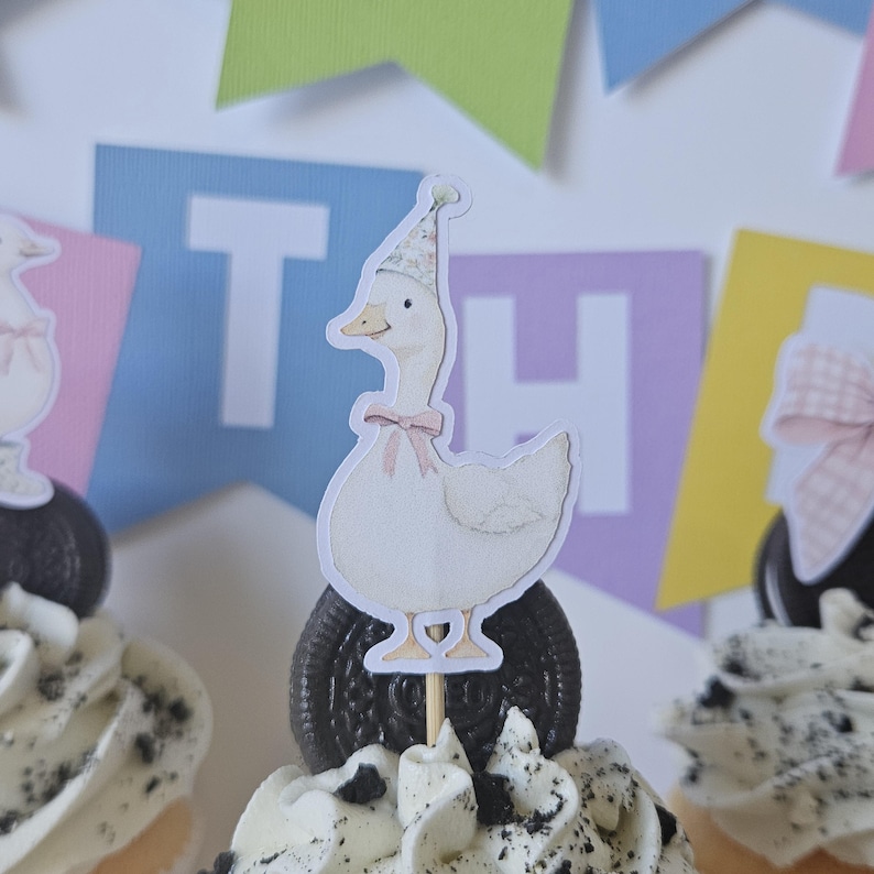 May include: A white goose cupcake topper featuring a floral party hat and a light pink bow around its neck, mounted on a dark chocolate sandwich cookie. The goose is illustrated in a soft style and is positioned atop a swirl of white frosting.