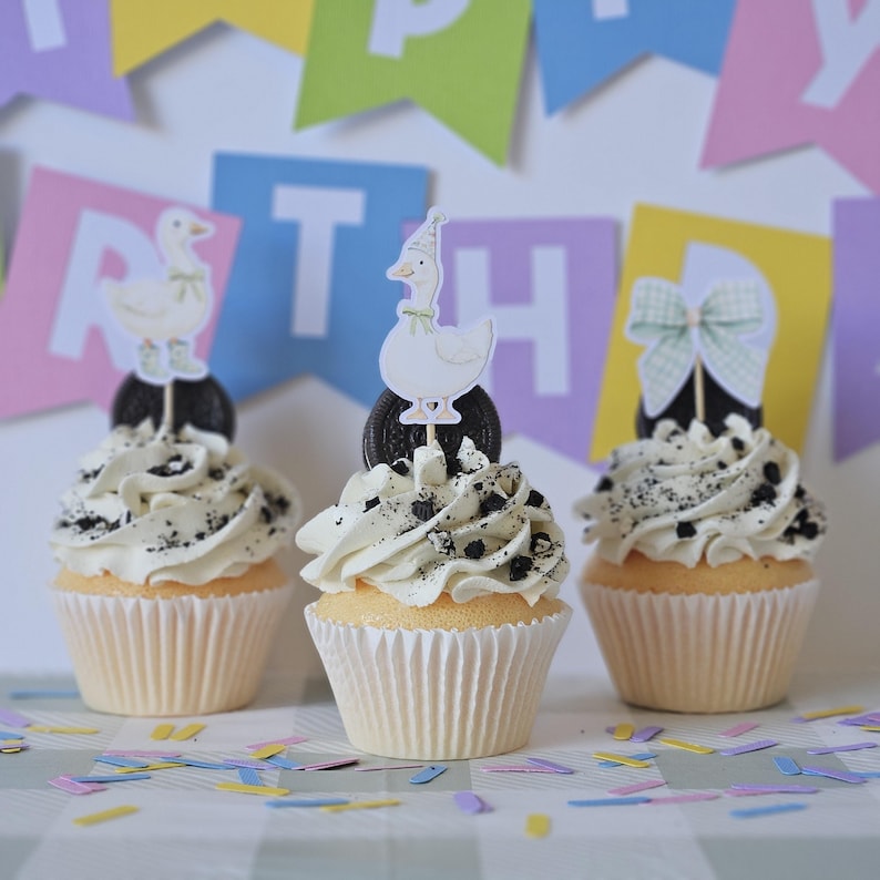 May include: Three cupcakes in white paper liners topped with white frosting and crushed dark cookie pieces. Two cupcakes feature a paper topper of a white duck wearing a party hat and green bow, while the third has a green gingham bow topper.