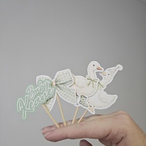 Puede incluir: Adornos para tartas de baby shower con un lazo verde y blanco, dos gansos y la frase "Baby Keane". Los gansos son blancos con detalles verdes y est&aacute;n sujetos a palitos de madera.