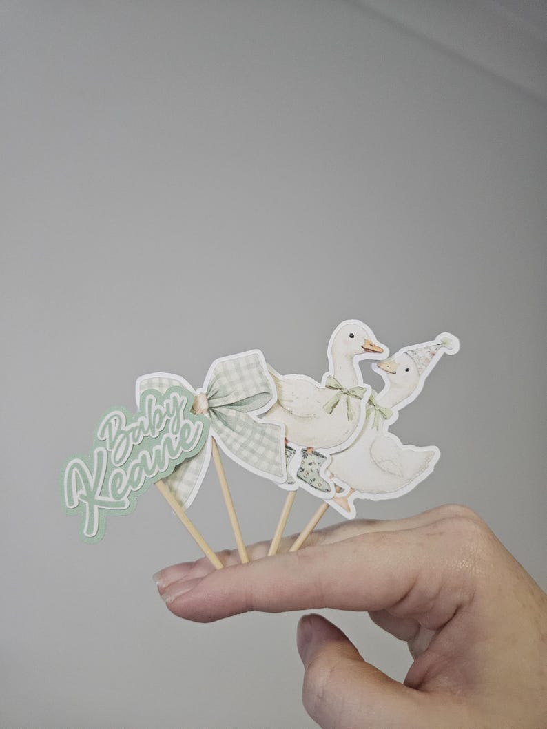 Puede incluir: Adornos para tartas de baby shower con un lazo verde y blanco, dos gansos y la frase "Baby Keane". Los gansos son blancos con detalles verdes y est&aacute;n sujetos a palitos de madera.