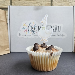 Puede incluir: Un cupcake con chispas de chocolate y un adorno de ganso. El cupcake est&aacute; en un envoltorio de papel blanco. En el fondo hay una caja con una pegatina que dice "CKCPTSAU Bringing Your Passion To Life".