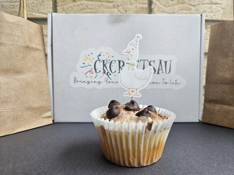Puede incluir: Un cupcake con chispas de chocolate y un adorno de ganso. El cupcake est&aacute; en un envoltorio de papel blanco. En el fondo hay una caja con una pegatina que dice "CKCPTSAU Bringing Your Passion To Life".
