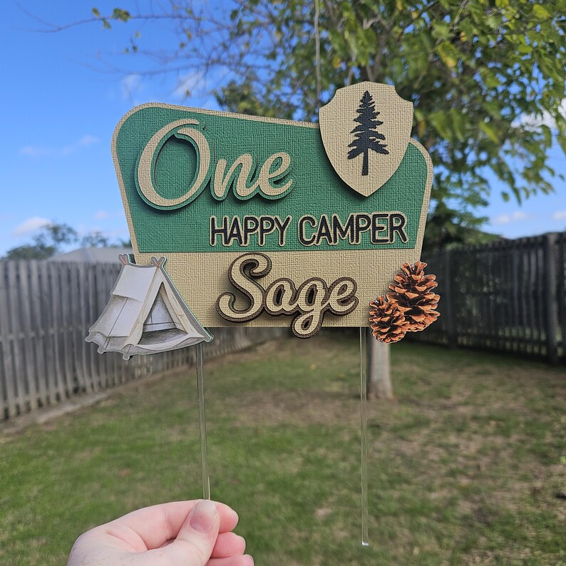 May include: A layered cake topper with the words "One HAPPY CAMPER Sage" in a camping theme. The topper includes a tent, pine tree, and pine cones. The background is green and tan.