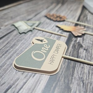 May include: A collection of decorative wooden skewers with camping-themed toppers. One skewer features a sign that reads "One HAPPY CAMPER" in a light green and beige color scheme. Other toppers include a tent and a pinecone.
