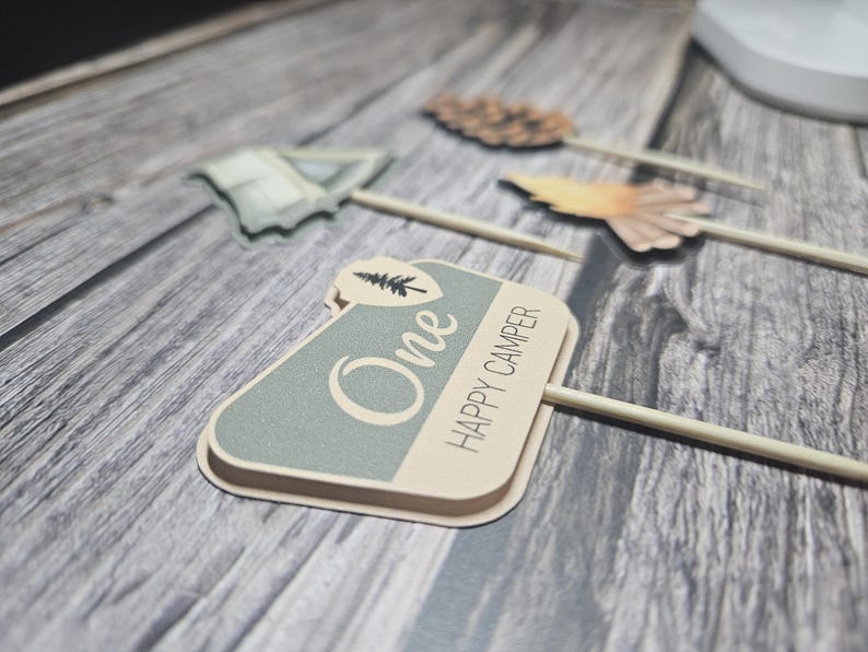 May include: A collection of decorative wooden skewers with camping-themed toppers. One skewer features a sign that reads "One HAPPY CAMPER" in a light green and beige color scheme. Other toppers include a tent and a pinecone.