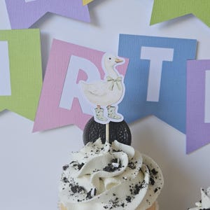 May include: A cupcake topper featuring a white duck illustration wearing a light green bow and matching floral patterned boots. The cutout is mounted on a wooden pick and inserted into a dark chocolate sandwich cookie atop a frosted cupcake.