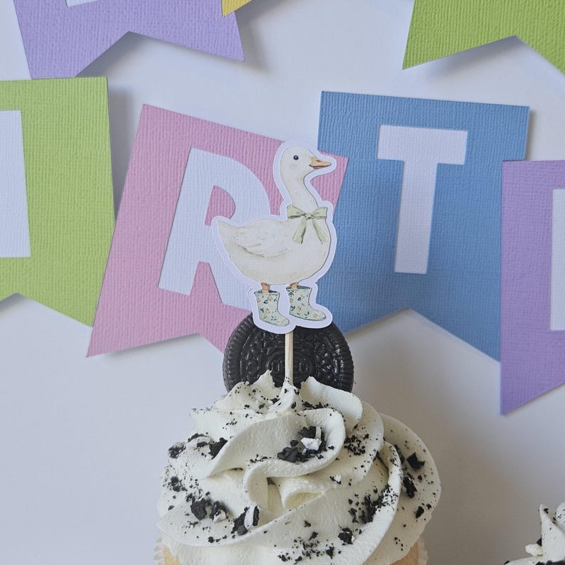 May include: A cupcake topper featuring a white duck illustration wearing a light green bow and matching floral patterned boots. The cutout is mounted on a wooden pick and inserted into a dark chocolate sandwich cookie atop a frosted cupcake.