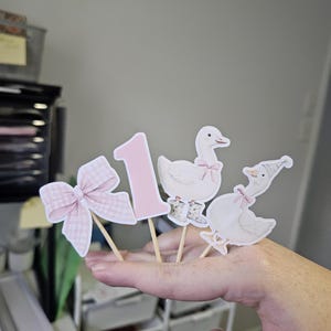 Puede incluir: Cuatro adornos decorativos para tartas sobre palitos de madera. Los adornos incluyen un lazo rosa a cuadros, el n&uacute;mero "1" y dos ilustraciones de gansos con lazos y sombreros. Los adornos son de color rosa claro y blanco.