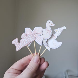 Puede incluir: Decoraciones de papel rosa y blanco sobre palitos de madera. El conjunto incluye un lazo con estampado de cuadros, el nombre "Lola" y dos gansos con lazos y sombreros. Probablemente se trate de adornos para tartas o decoraciones para fiestas.