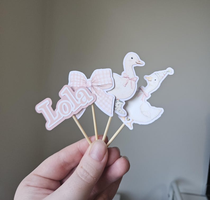 Puede incluir: Decoraciones de papel rosa y blanco sobre palitos de madera. El conjunto incluye un lazo con estampado de cuadros, el nombre "Lola" y dos gansos con lazos y sombreros. Probablemente se trate de adornos para tartas o decoraciones para fiestas.
