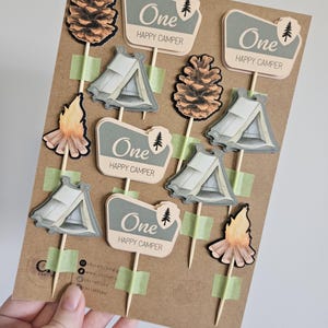May include: A collection of camping-themed cupcake toppers. The toppers include images of tents, pinecones, and campfires. Each topper has the text "One HAPPY CAMPER" on a sign. The toppers are attached to wooden sticks with green tape.