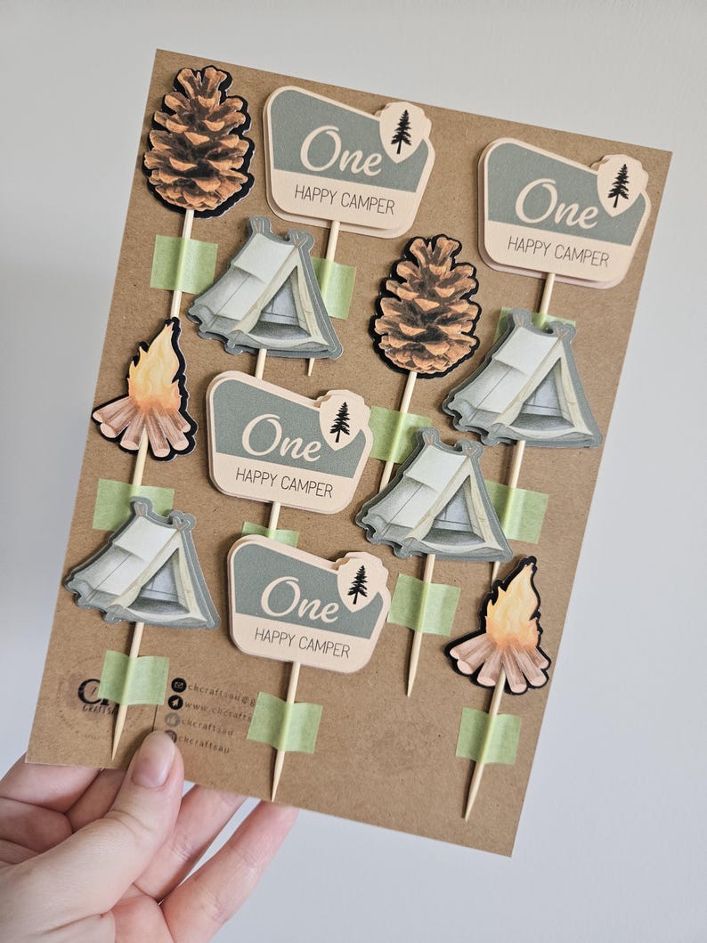 May include: A collection of camping-themed cupcake toppers. The toppers include images of tents, pinecones, and campfires. Each topper has the text "One HAPPY CAMPER" on a sign. The toppers are attached to wooden sticks with green tape.
