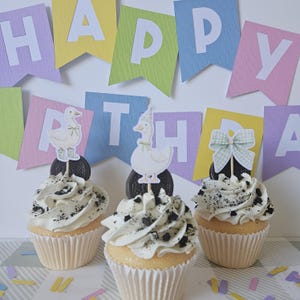 May include: Three cupcakes with white frosting and dark cookie crumbles, each topped with a dark sandwich cookie and a paper cutout. The cutouts feature a white goose in boots, a goose in a party hat, and a light green gingham patterned bow.