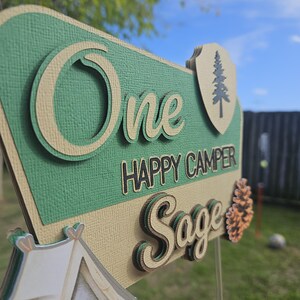 May include: A layered sign with the words "One Happy Camper Sage" in a camping theme. The sign is green, tan, and brown with a tent and pinecone accent. The background is a grassy yard with a blue sky.