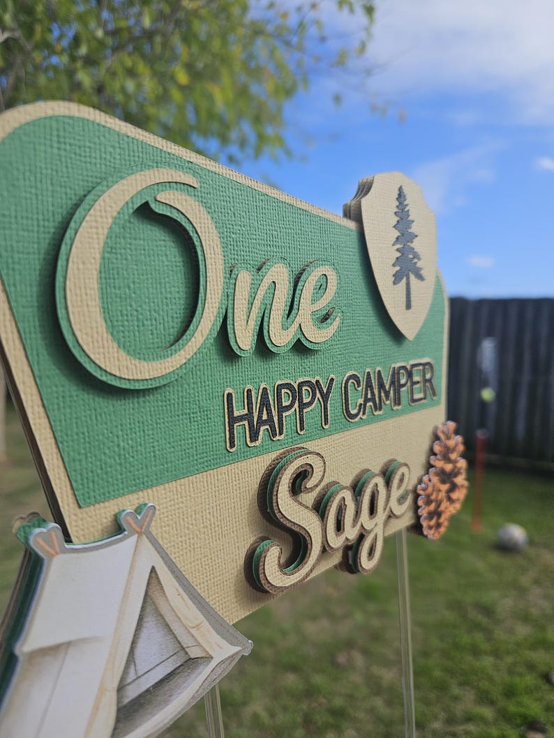 May include: A layered sign with the words "One Happy Camper Sage" in a camping theme. The sign is green, tan, and brown with a tent and pinecone accent. The background is a grassy yard with a blue sky.
