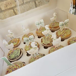 Puede incluir: Una caja de panader&iacute;a blanca llena de cupcakes decorados. Cada cupcake est&aacute; adornado con glaseado verde, chispas y un adorno en forma de pato. Algunos cupcakes tienen adornos en forma de lazo. Tambi&eacute;n se ve un adorno con el n&uacute;mero 1.