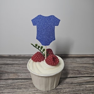 Op de afbeelding: Een cupcake met witte frosting, gegarneerd met rode frambozen en een blauwe glitter romper decoratie. De cupcake staat in een witte papieren cup, op een houten achtergrond.