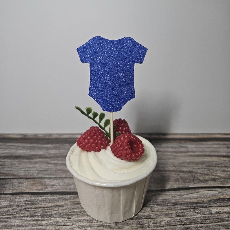 Op de afbeelding: Een cupcake met witte frosting, gegarneerd met rode frambozen en een blauwe glitter romper decoratie. De cupcake staat in een witte papieren cup, op een houten achtergrond.