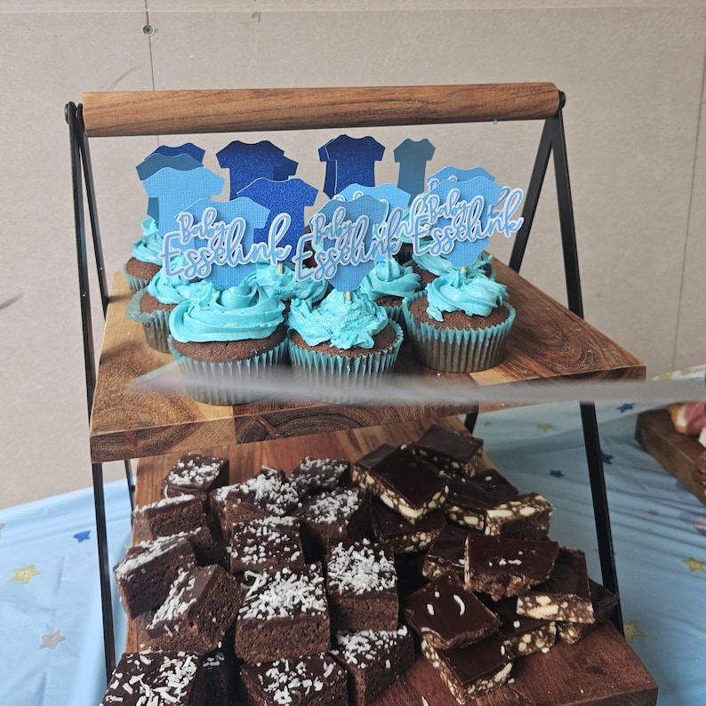 Op de afbeelding: Een houten serveerstandaard met twee niveaus toont cupcakes met blauwe frosting en toppers met de tekst "Baby Esselnik". De bovenste laag bevat cupcakes, terwijl de onderste laag brownies en chocoladeblokjes bevat. De standaard heeft een zwart metalen frame.