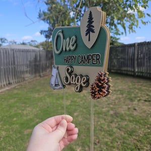 May include: A layered wooden cake topper with the words "One Happy Camper Sage". The topper features a tent, pine cone, and a pine tree design. The topper is held by a clear plastic stick.