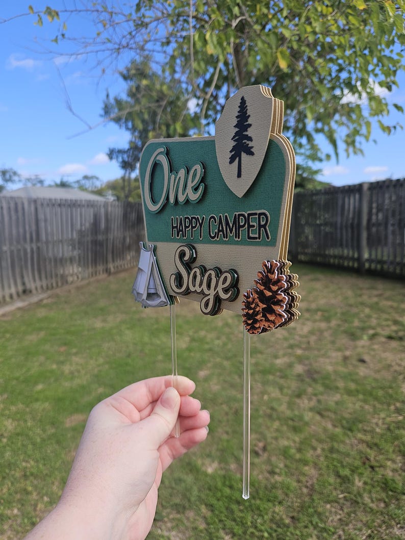 May include: A layered wooden cake topper with the words "One Happy Camper Sage". The topper features a tent, pine cone, and a pine tree design. The topper is held by a clear plastic stick.
