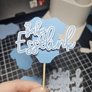 Op de afbeelding: Een lichtblauwe papieren uitsparing in de vorm van een babyromper met de woorden "Baby Esselink" in witte letters. De uitsparing is bevestigd aan een houten stokje, waarschijnlijk een taarttopper. Andere blauwe romperuitsparingen zijn zichtbaar.