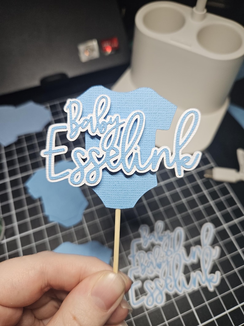 Op de afbeelding: Een lichtblauwe papieren uitsparing in de vorm van een babyromper met de woorden "Baby Esselink" in witte letters. De uitsparing is bevestigd aan een houten stokje, waarschijnlijk een taarttopper. Andere blauwe romperuitsparingen zijn zichtbaar.