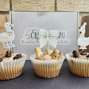 Puede incluir: Tres cupcakes con adornos decorativos. Dos cupcakes tienen adornos de ganso, uno con botas. El cupcake central tiene un adorno de lazo y galletas peque&ntilde;as con forma de animal. Todos los cupcakes est&aacute;n en moldes de papel blancos y cubiertos con chispas de chocolate.