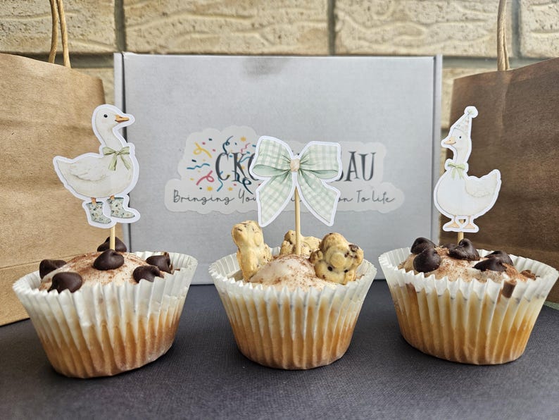 Puede incluir: Tres cupcakes con adornos decorativos. Dos cupcakes tienen adornos de ganso, uno con botas. El cupcake central tiene un adorno de lazo y galletas peque&ntilde;as con forma de animal. Todos los cupcakes est&aacute;n en moldes de papel blancos y cubiertos con chispas de chocolate.