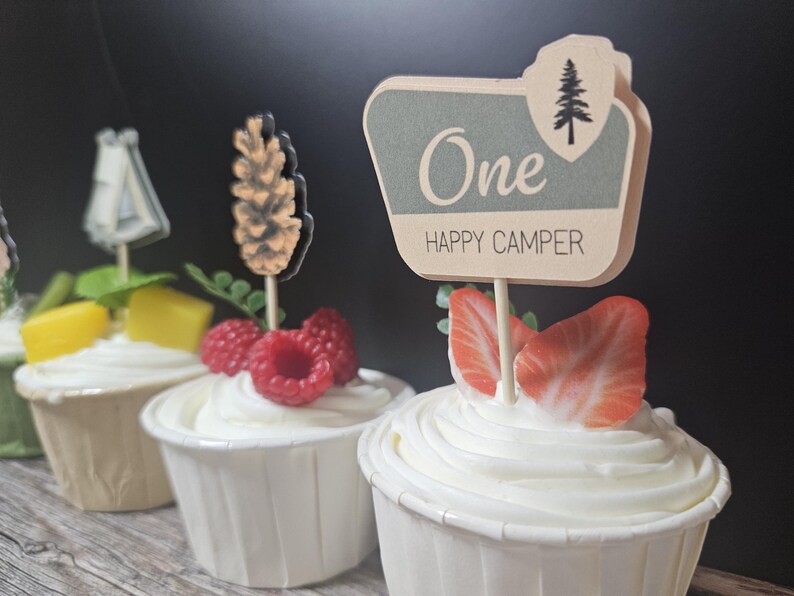 May include: Cupcakes with white frosting, topped with fruit and decorative toppers. One cupcake has a sign that reads "One HAPPY CAMPER" with a tree graphic. Other toppers include a pinecone and a tent. The cupcakes are in white paper cups.